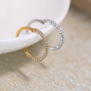 May include: Two circular hoop earrings, one in a gold tone and one in a silver tone, each featuring a row of small, clear, sparkling stones embedded along the outer edge of the hoop. The earrings are thin, metallic, and circular in shape.
