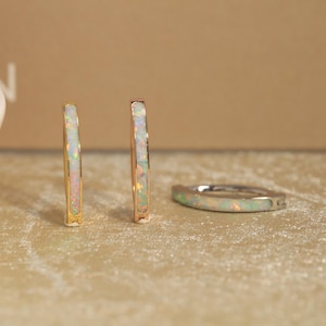 May include: Two upright hoop earrings and one lying flat hoop earring, all featuring a metallic band inlaid with iridescent white opal stones that display flashes of green, pink, and blue color throughout the surface of each piece.