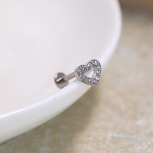 Puede incluir: Una pieza de joyería corporal tipo labret en tono plateado con un adorno en forma de corazón. El contorno del corazón está incrustado con pequeñas piedras transparentes y brillantes en su parte frontal. El poste es recto y termina en un disco plano.