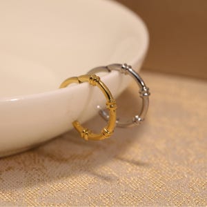 May include: Two hoop earrings, one gold-toned and one silver-toned, are displayed. The earrings have a bamboo-like design with segmented details. They are placed near a white, curved surface, set against a textured, golden background.
