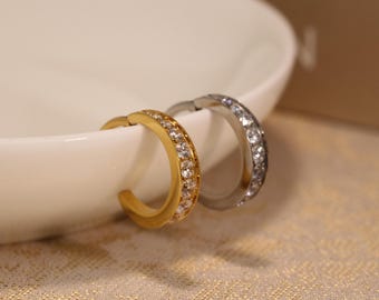 May include: A close-up shot of two hoop earrings. One is gold-coloured, the other is silver-coloured. Both are adorned with small, clear gemstones. The earrings are placed on a white, curved surface, against a soft, patterned background.
