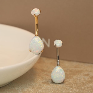 Puede incluir: Dos anillos de ombligo de oro con piedras de ópalo blanco en forma de lágrima. Los anillos están sobre una superficie blanca.