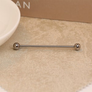 Op de afbeelding: Een zilveren industriële barbell body piercing. De barbell heeft een rechte, slanke staaf met aan elk uiteinde een kleine, ronde bal. De sieraden worden gepresenteerd op een beige oppervlak.