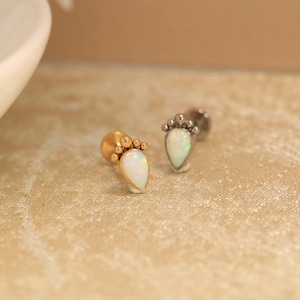 May include: Two stud earrings featuring teardrop-shaped white opals set in metal. One earring is gold-toned and the other is silver-toned. Each opal is accented with a decorative fan of small metal spheres along the top edge of the setting.
