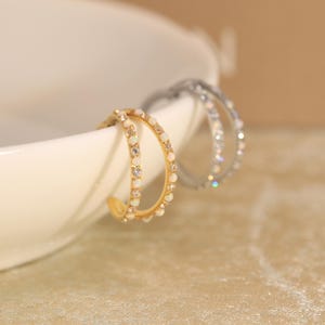May include: Two open hoop earrings, one in gold tone and one in silver tone, each featuring a double-band design. The bands are embellished with alternating small, round, iridescent white opal-like stones and clear, sparkling, faceted crystals.
