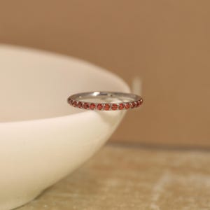 May include: A thin silver-toned metal band ring featuring a continuous row of small, round, vibrant orange-red gemstones set along the outer circumference. The ring has a polished metallic finish and a slim, uniform profile throughout the design.
