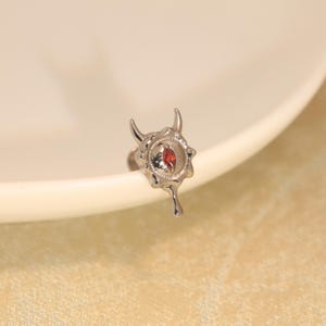May include: A silver-toned metal body jewelry stud featuring a stylized devil horn design with a central marquise-cut red gemstone. The piece has a molten, irregular edge texture and a small drip-like extension at the bottom of the setting.