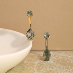 Könnte beinhalten: Zwei Bauchnabelpiercings mit tropfenförmigen Moosachat-Steinen mit grünen Einschlüssen. Ein Piercing hat einen goldfarbenen Metallstab, das andere einen silberfarbenen Metallstab. Beide haben eine kleine, runde Moosachat-Kugel als Verschluss.