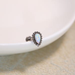 May include: A teardrop-shaped body jewellery stud featuring a central iridescent white opal stone surrounded by a decorative border of small dark metal beads. The piece includes a flat circular back and a short post for secure placement.