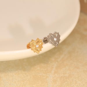 Può includere: Due orecchini a lobo a forma di cuore, uno color oro e uno color argento, ciascuno caratterizzato da un motivo inciso a raggiera che si irradia da una gemma centrale trasparente. Gli orecchini presentano un bordo testurizzato e una chiusura a perno.