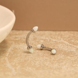 May include: Two curved barbell piercings featuring a metallic silver-toned finish. Each end of the curved bars is capped with a smooth, rounded white opal-like stone. The jewelry pieces are positioned with one standing upright and one lying flat.