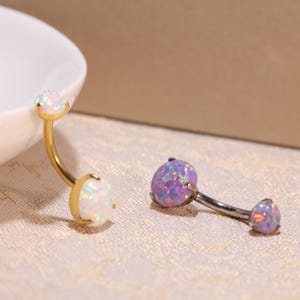 Könnte beinhalten: Zwei Bauchnabelringe mit Opal-Steinen. Ein Ring hat einen weißen Opal-Stein und der andere einen lila Opal-Stein.
