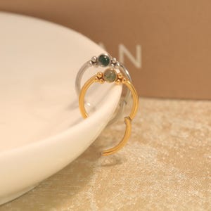 May include: Two circular hinged hoop rings, one in silver tone and one in gold tone, each featuring a small round green stone set at the top with decorative metal bead accents on either side of the stone, designed for body piercing jewelry.