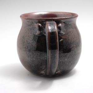 May include: A dark brown ceramic mug with a speckled glaze. The mug has a rounded body and a thick, rounded handle.