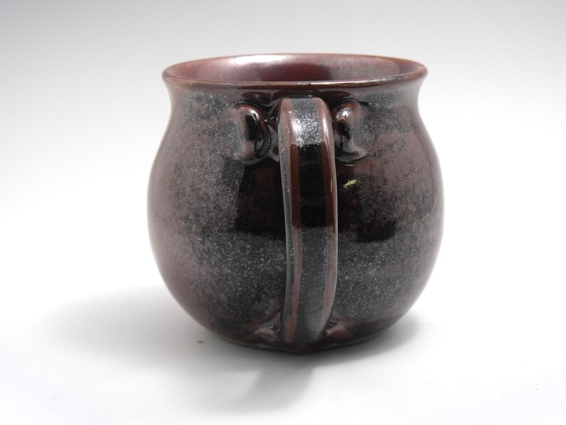 May include: A dark brown ceramic mug with a speckled glaze. The mug has a rounded body and a thick, rounded handle.