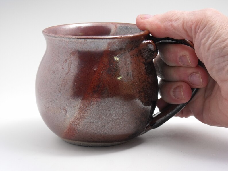 May include: A brown and white ceramic mug with a rounded body and a black handle. The mug has a speckled glaze and a slightly uneven surface.