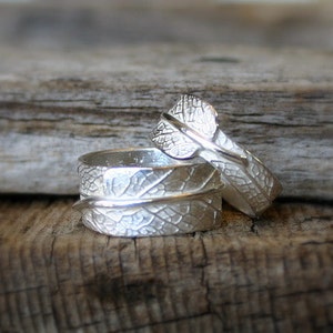 Puede incluir: Dos anillos de plata con un diseño de hoja. Los anillos están apilados uno encima del otro y están sobre un trozo de madera flotante.