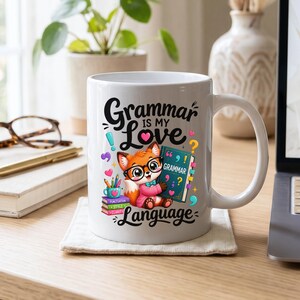 Puede incluir: Una taza de cerámica blanca con un zorro de dibujos animados que lleva gafas, sostiene un libro titulado Grammar y está rodeado de material escolar colorido. El texto dice Grammar is my Love Language, con signos de puntuación y corazones.