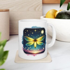 May include: White ceramic mug with a colorful illustration of a glowing firefly against a starry night sky. The firefly has yellow and green wings and a bright yellow body. The background is a watercolor wash of blue, purple, and pink.