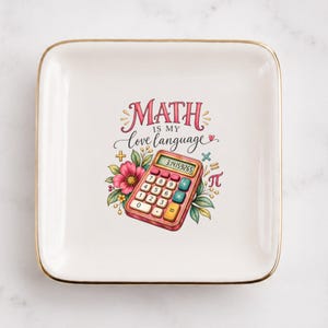 May include: A square white ceramic trinket dish with a gold-toned rim. The center features a colorful illustration of a calculator surrounded by flowers and the text "MATH IS MY love language" with mathematical symbols including a pi symbol.