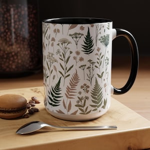 May include: A white ceramic mug with a black handle and rim. The mug features a botanical pattern with green and brown ferns and flowers on a white background.