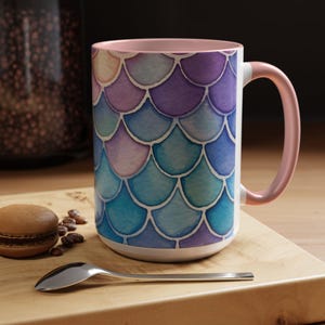 May include: A white ceramic mug with a pink rim and handle. The mug features a watercolor design of blue, purple, and pink scales.