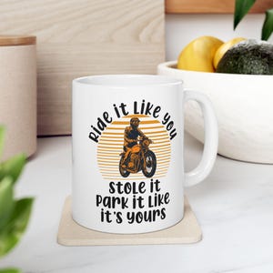 May include: White ceramic mug with a black and orange graphic of a person riding a motorcycle. The text on the mug reads "Ride it like you stole it park it like it's yours".