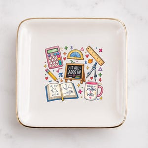 May include: A square white ceramic trinket dish with a gold-toned rim featuring a colorful illustration of math-themed items including a calculator, protractor, ruler, compass, an open textbook, and a mug with the text "FUELED BY COFFEE & MATH".