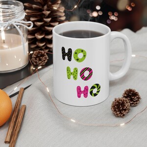 Könnte beinhalten: Eine weiße Keramiktasse mit einem schwarzen, grünen und rosa "HO HO HO"-Design. Die Tasse ist mit Kaffee gefüllt und steht auf einem Tisch mit einer weißen Tischdecke.