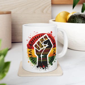 Peut inclure: Une tasse en céramique blanche avec un graphique coloré d'un poing levé avec un motif noir et rouge. Le poing est entouré d'un cercle rouge, jaune et vert avec un effet de peinture éclaboussé.