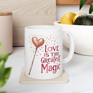 May include: White ceramic mug with a pink heart-shaped wand with gold glitter and the text "Love is the greatest magic" in pink.