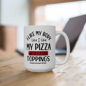 May include: White ceramic mug with the text "I like my body like I like my pizza with extra toppings" in black and red. The mug has a large handle and is sitting on a wooden surface.