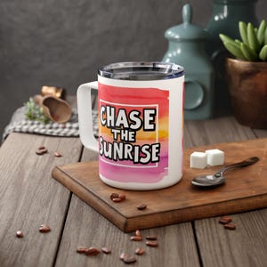 May include: White travel mug with a pink, orange and yellow watercolour sunset design. The mug has a black handle and the text "CHASE THE SUNRISE" in a black box.