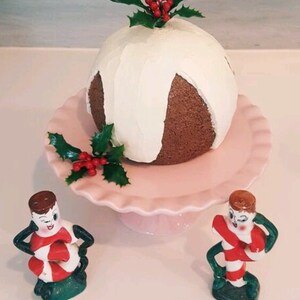 May include: A white and brown Christmas cake with white frosting and holly on top, sitting on a pink cake stand. Two candy cane figurines are on either side of the cake stand.