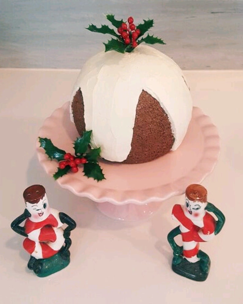 May include: A white and brown Christmas cake with white frosting and holly on top, sitting on a pink cake stand. Two candy cane figurines are on either side of the cake stand.