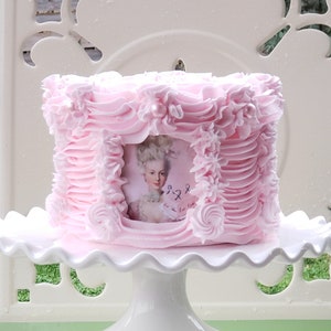 May include: A pink cake decorated with pink frosting and pearl accents. The cake has a framed image of a woman in a white dress and a white wig. The cake is on a white cake stand.