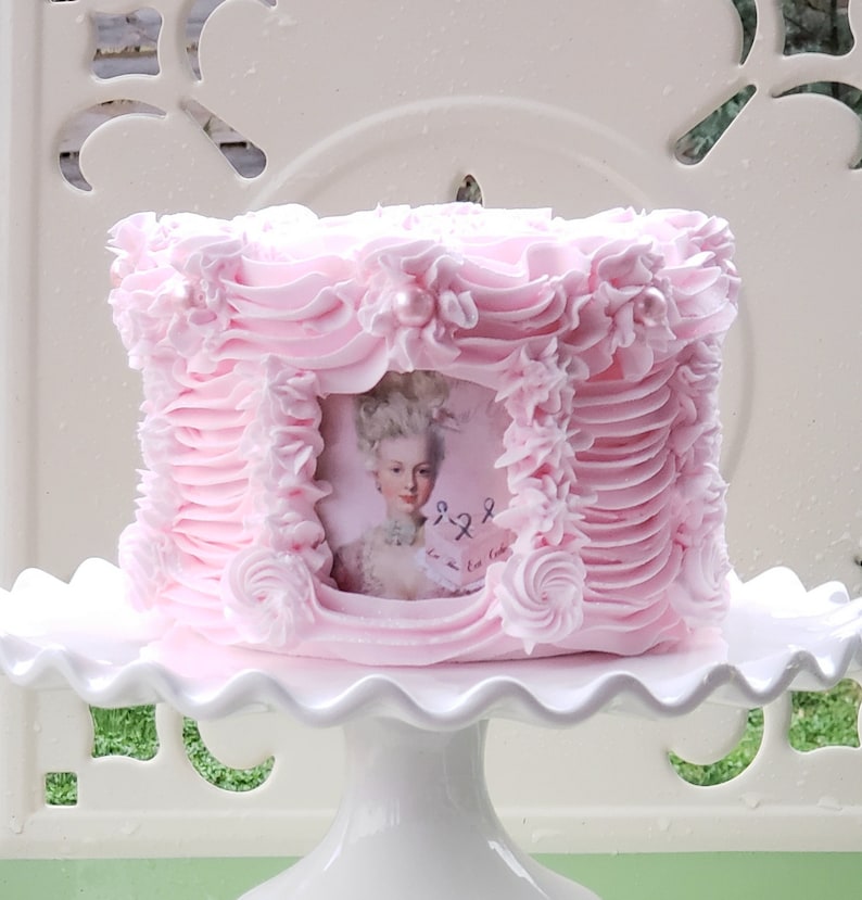 May include: A pink cake decorated with pink frosting and pearl accents. The cake has a framed image of a woman in a white dress and a white wig. The cake is on a white cake stand.