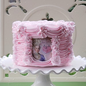 May include: A pink cake decorated with pink frosting and pearl accents. The cake has a framed image of a woman in a vintage dress. The cake is on a white cake stand.
