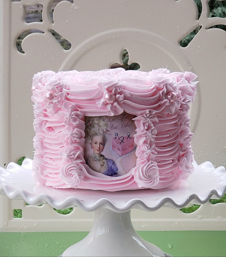 May include: A pink cake decorated with pink frosting and pearl accents. The cake has a framed image of a woman in a vintage dress. The cake is on a white cake stand.
