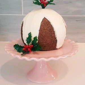 May include: A white and brown Christmas pudding with holly and red berries on top, displayed on a pink cake stand.