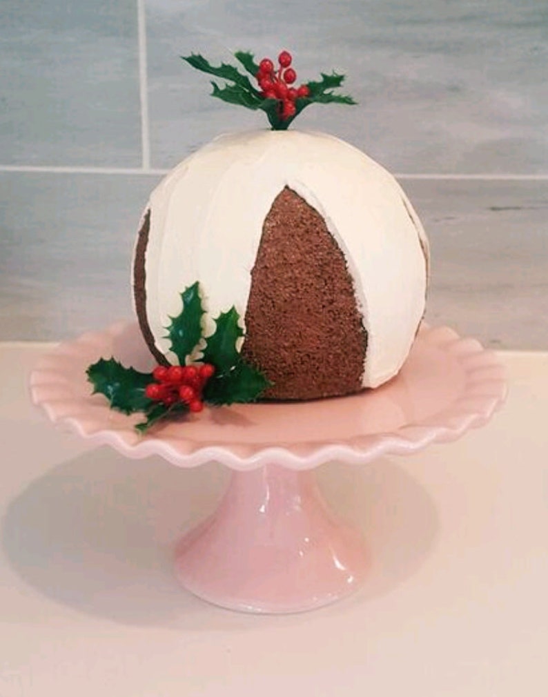 May include: A white and brown Christmas pudding with holly and red berries on top, displayed on a pink cake stand.
