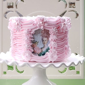May include: A pink cake decorated with pink frosting and a vintage image of a woman in a dress. The cake is on a white cake stand.