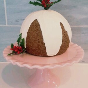 May include: A white and brown Christmas pudding with a white icing drizzle, topped with a sprig of holly with red berries, sitting on a pink cake stand.