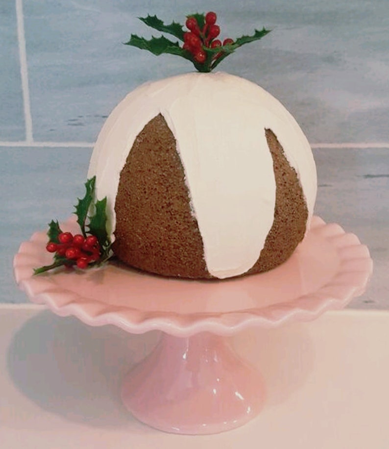 May include: A white and brown Christmas pudding with a white icing drizzle, topped with a sprig of holly with red berries, sitting on a pink cake stand.