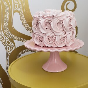 Puede incluir: Un pastel rosa decorado con rosas de glaseado se encuentra sobre un soporte para pastel rosa. El soporte para pastel est&aacute; sobre una mesa amarilla.