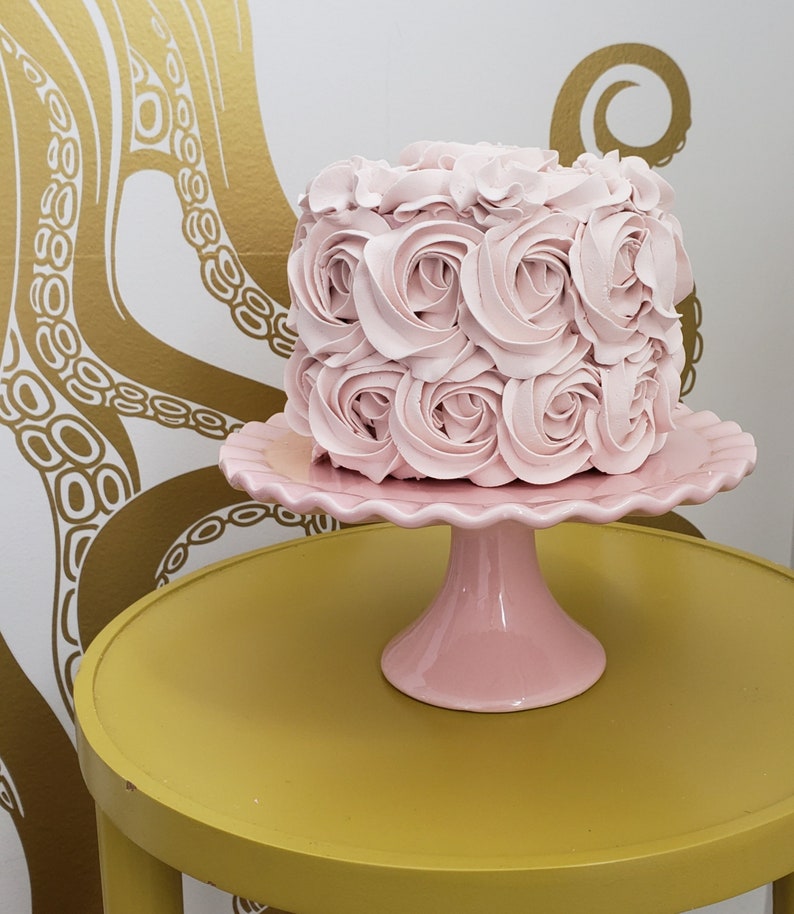 Puede incluir: Un pastel rosa decorado con rosas de glaseado se encuentra sobre un soporte para pastel rosa. El soporte para pastel est&aacute; sobre una mesa amarilla.