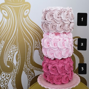 Puede incluir: Un pastel de tres pisos decorado con rosas de glaseado rosa, rosa claro y rosa p&aacute;lido. El pastel est&aacute; sobre un soporte para pastel rosa con un borde festoneado.