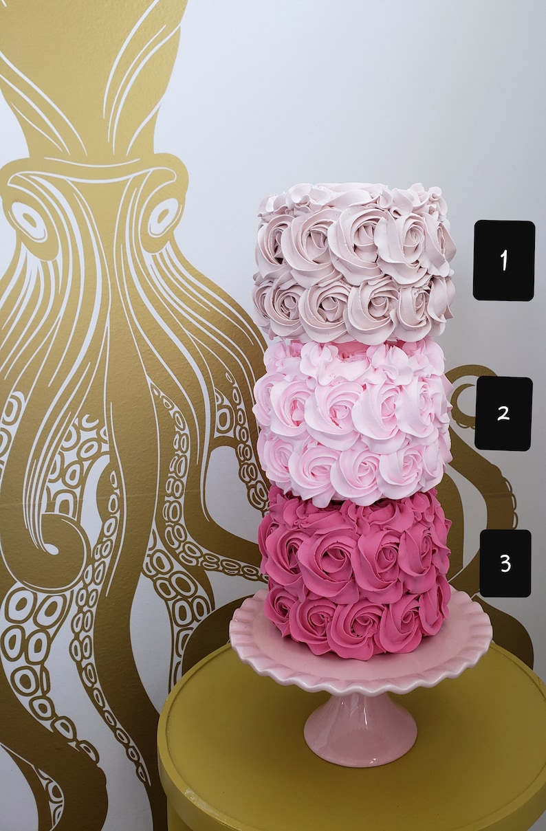Puede incluir: Un pastel de tres pisos decorado con rosas de glaseado rosa, rosa claro y rosa p&aacute;lido. El pastel est&aacute; sobre un soporte para pastel rosa con un borde festoneado.