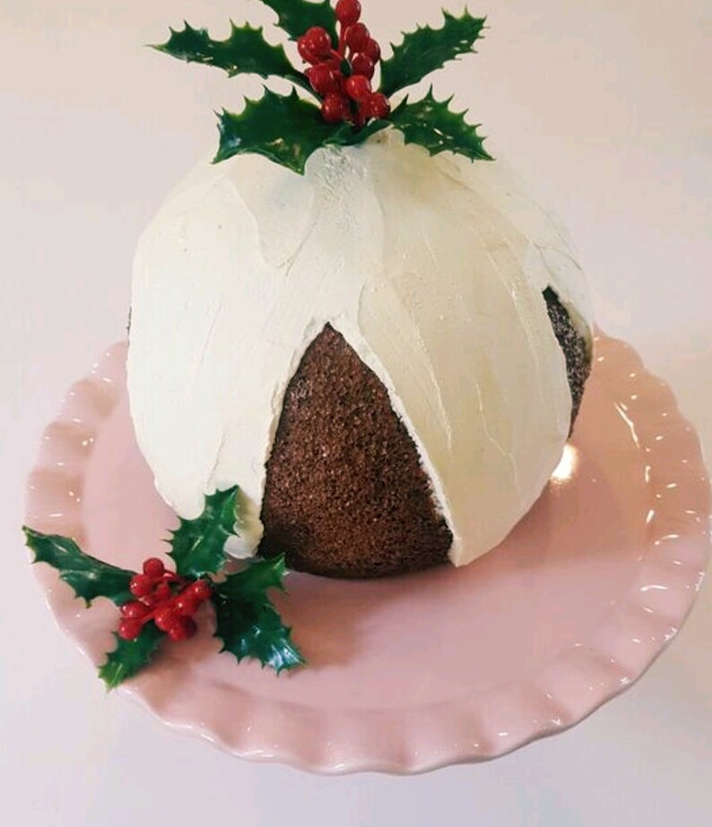 May include: A chocolate cake decorated with white frosting and holly sprigs with red berries. The cake is on a pink cake stand.