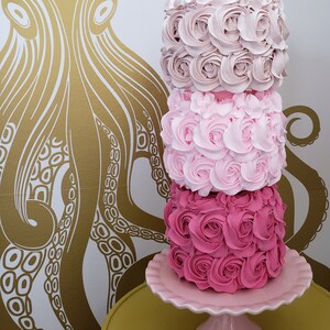 Puede incluir: Un pastel de tres pisos decorado con glaseado rosa y rosa claro, que se asemeja a rosas. El pastel est&aacute; sobre un soporte para pastel rosa con un borde festoneado. El soporte para pastel est&aacute; sobre una mesa amarilla.